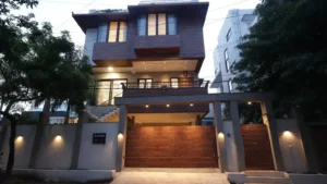 Modern three-story house at dusk with illuminated windows, a wooden gate, concrete pillars, a balcony with railings, an external staircase on the left side, and trees flanking the property.
