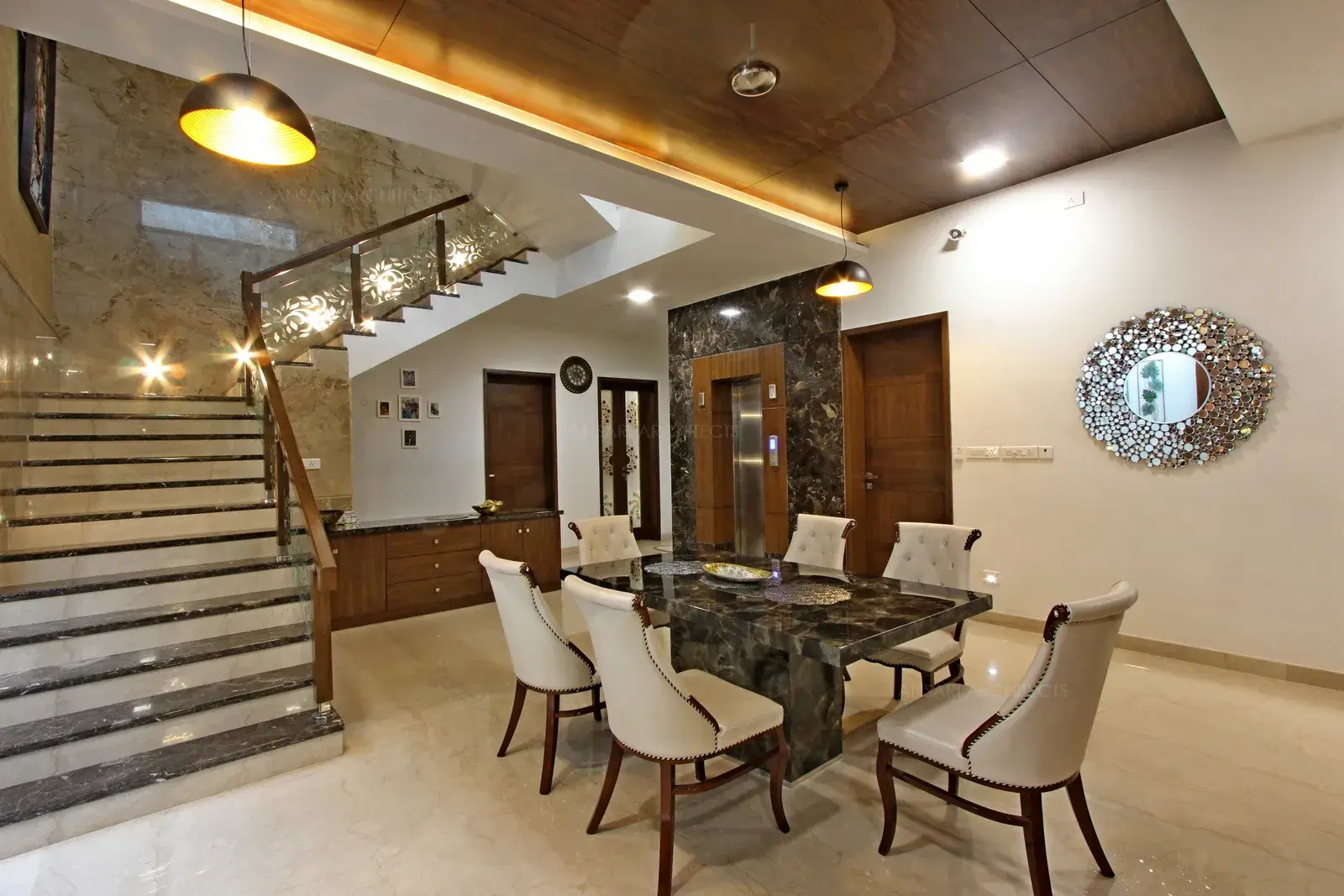 An ornate silver element with mirror on the wall and the cladding around lift highlights look of an understated dining space. Recessed cove lighting infuses warmth and imparts brightness into the dining space. The ceiling demarcates the dining area from rest of the space.