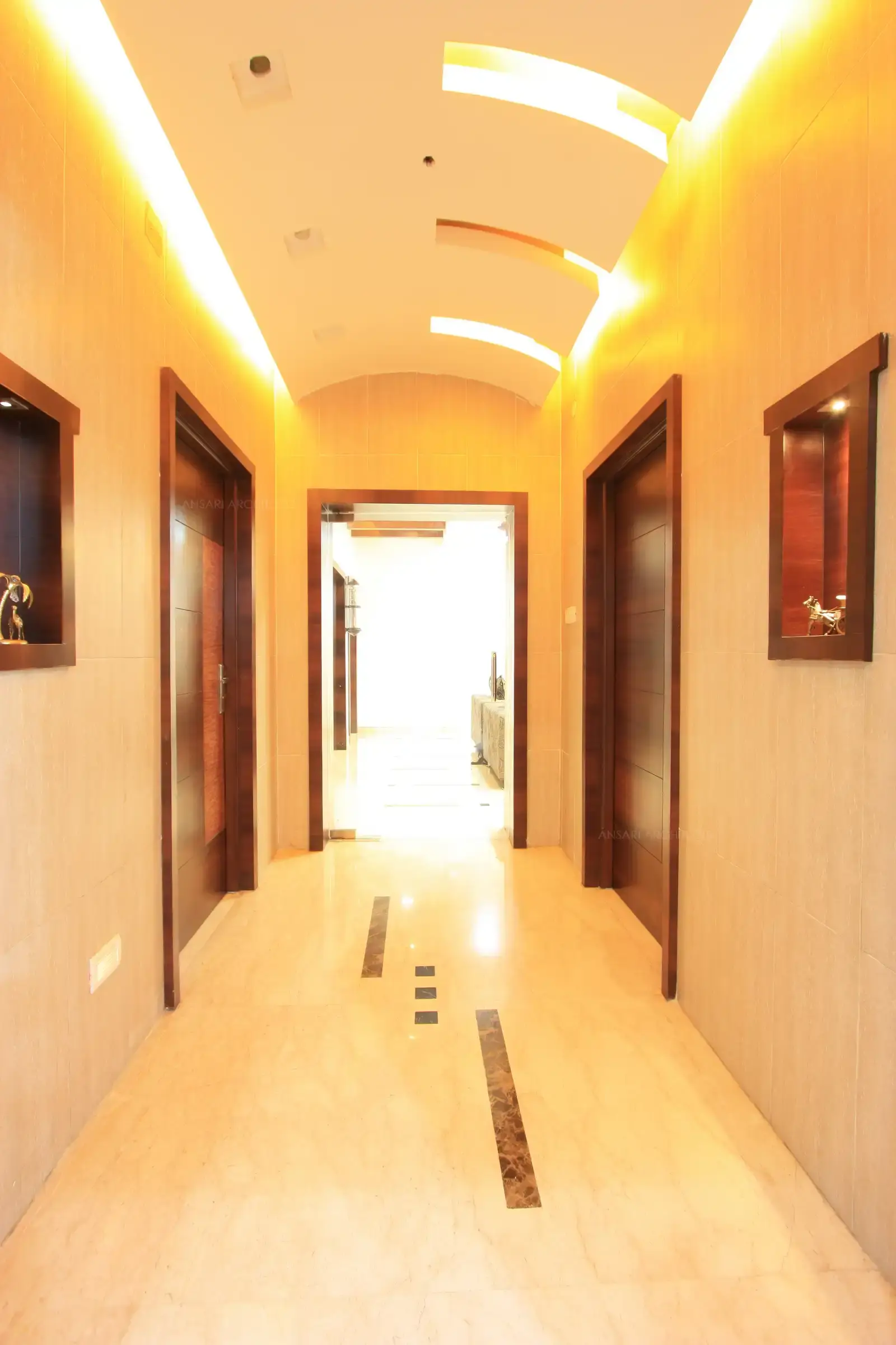 Wide hallway with a high ceiling, wooden accents, and elegant lighting. Features two doors and decorative wall niches. Bright, inviting space leading to a room in the distance.
