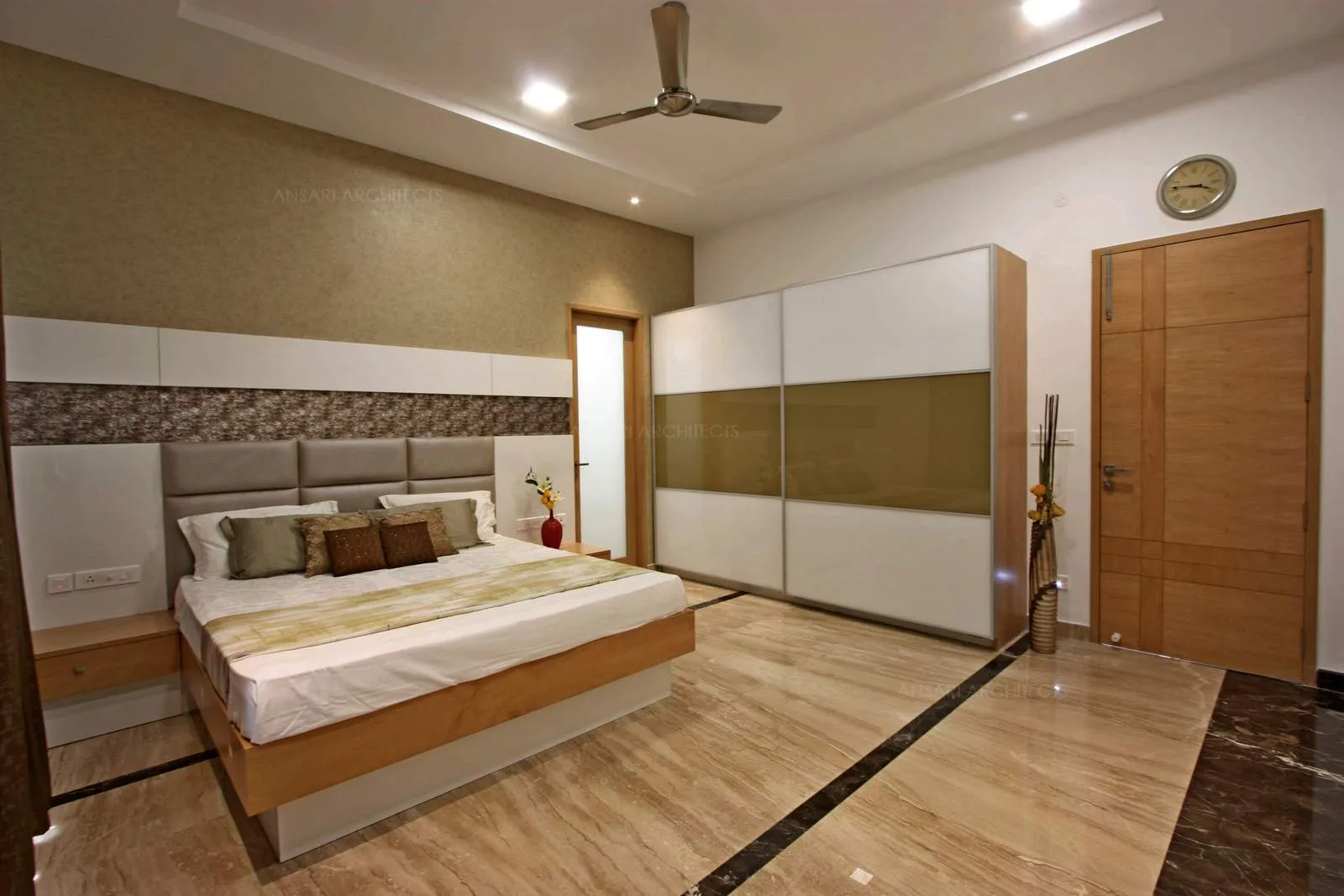 The bedroom is tactile with variety of textures, the wallpaper, the headboard panel with cushion tufted in the centremalong with wooden side table, and soft wooden finished wardrobe.