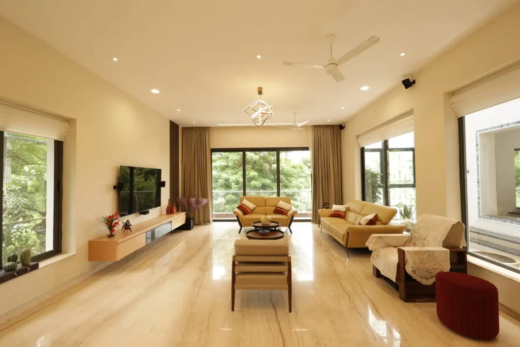Spacious living room with cream-colored walls and floor, large windows with beige curtains, two beige sofas with accent pillows, a beige armchair with lace covers, a red ottoman, a wall-mounted flat-screen TV, and a modern geometric ceiling light fixture.