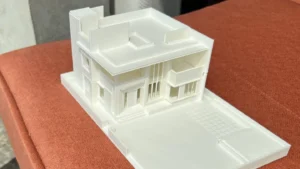 White architectural model of a modern two-story house with a flat roof, balconies, and a front entrance, placed on an orange textured surface.