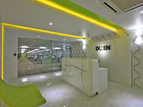 Modern reception area with white geometric desk, illuminated yellow ceiling strip lighting, green chair, and glass doors leading to a large office inside