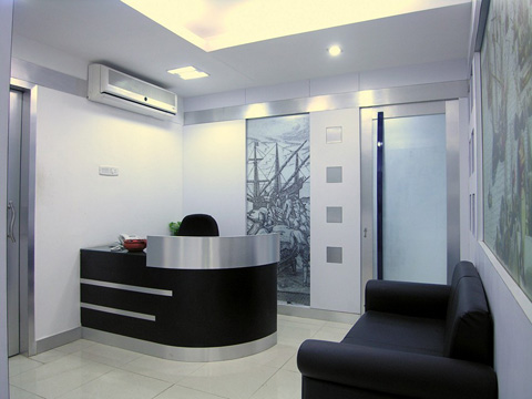 Modern office reception area with a curved black and silver desk, black chair behind the desk, black sofa to the right, artwork on the walls, white tiled floor, and air conditioning unit above the desk