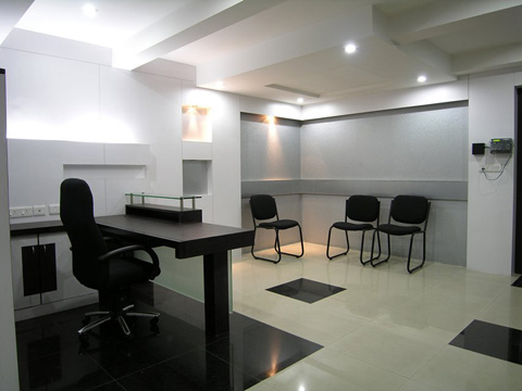 Modern office reception area with a black desk and swivel chair on the left, three black visitor chairs against a light gray wall on the right, and glossy tiled floors with black and beige patterns under bright ceiling lights.