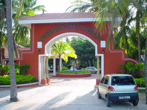 Entrance archway of Green coconut Resort with red walls and a tiled roof, surrounded by palm trees and greenery, with a small car parked on the right side of the driveway.