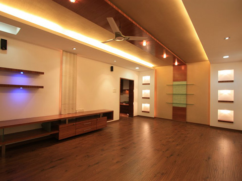 A modern empty living room with wooden flooring, built-in shelves with recessed lighting, a ceiling fan, and warm ambient lighting on the ceiling.