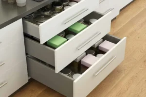 Open white kitchen drawers filled with organized jars and containers, some with pastel-colored lids, set against a wooden floor and a gray countertop.